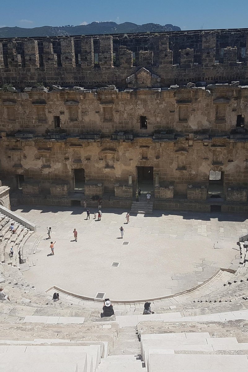 The towering stage wall of the Roman theatre at Aspendos rising above the sweeping semicircle of stone seating, with visitors small in the orchestra below and the Taurus foothills beyond under a clear sky. Serik, Antalya, Türkiye.